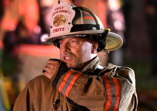 Photo of Fire Chief Fritz at an incident. Wearing white chiefs helmet, talking on the radio.
